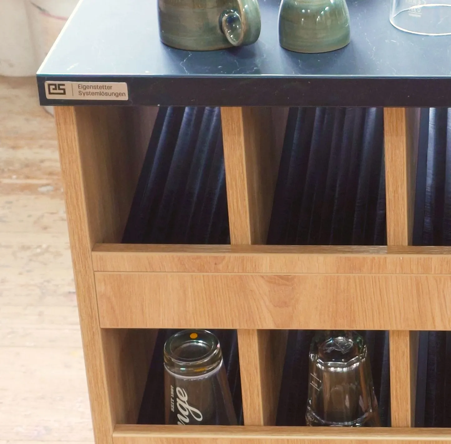 Servicemöbel aus hellem Holz mit dunkler Steinplatte und Eigenstetter-Logoplakette, darauf Keramikbecher und ein Glas, im Korpus stoffgefütterte Fächer mit Gläsern und Flaschen